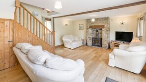 A spacious living room with a stone fireplace, white sofas, a wooden staircase, and a television. - Groves Fishleigh (Okehampton)