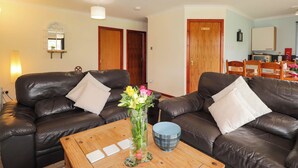 A living room with two black leather sofas, a wooden coffee table, and a vase with flowers. There are wooden doors and a small table with a lamp in the background.