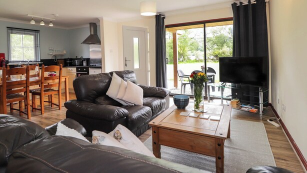 A living room with a leather sofa, wooden coffee table, and a TV on a stand. There is a kitchen in the background with a stove and a window.