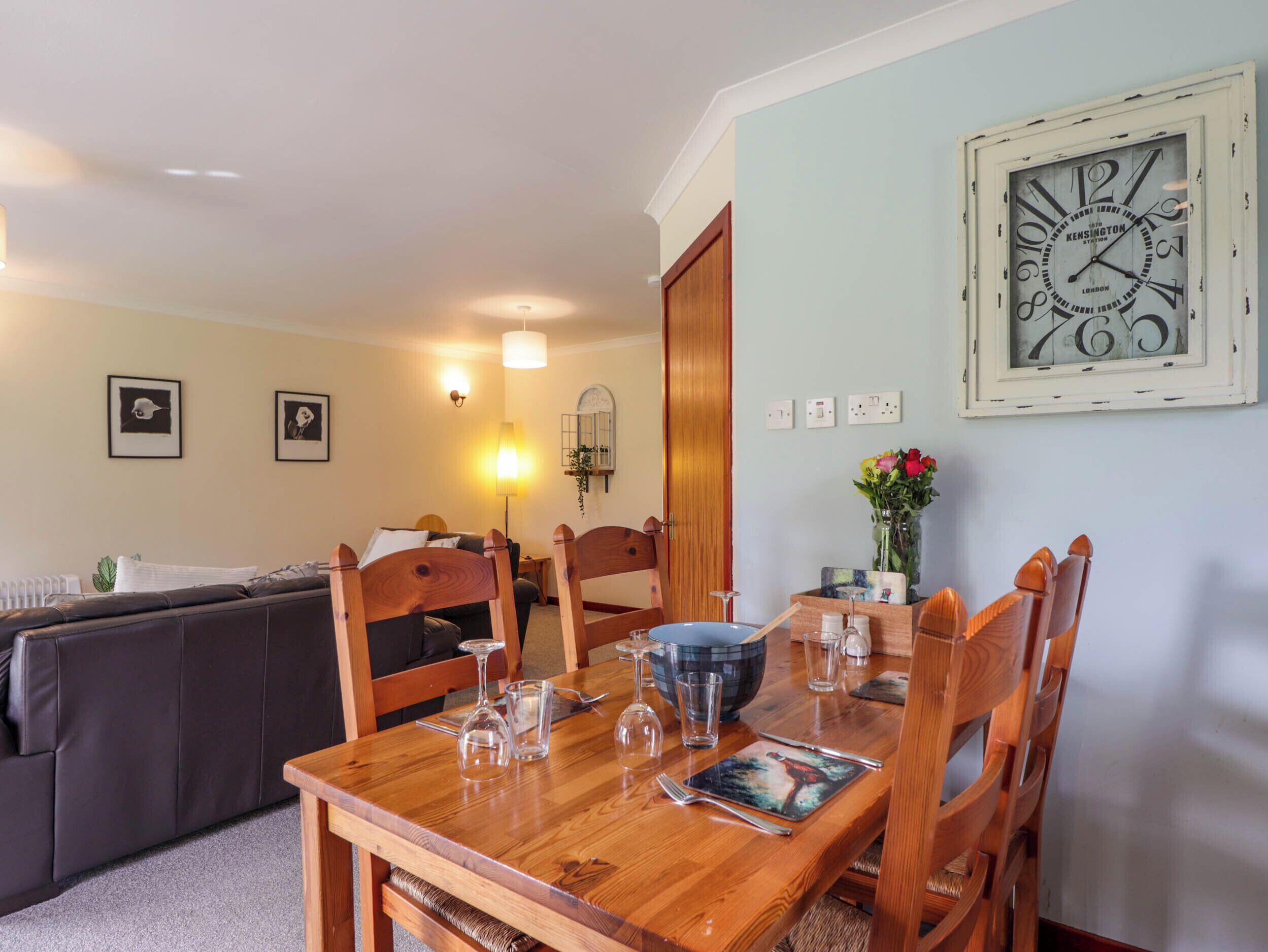 A dining area with a wooden table set for four, surrounded by wooden chairs. A large wall clock is visible on the wall.