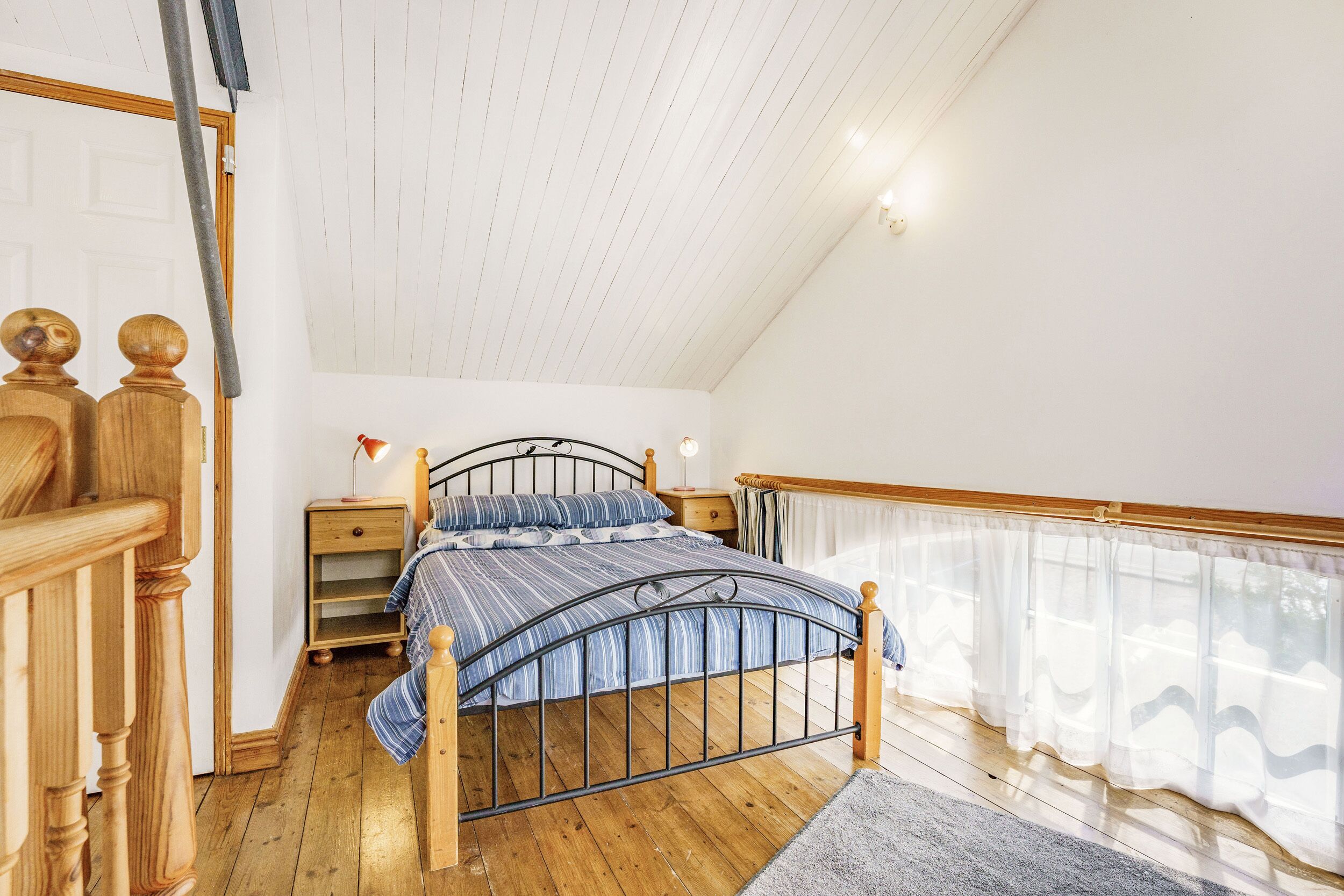 A bedroom with a wooden bed frame, a nightstand, and a window with sheer curtains.