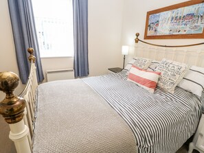 A neatly made bed with striped bedding and decorative pillows, a bedside table with a lamp, and a window with curtains. - Driftwood Apartment (Morpeth)