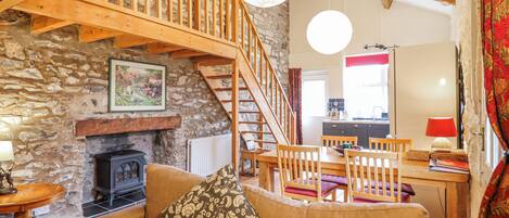A cozy living room with a stone fireplace, wooden staircase, and a dining area with chairs and a table.