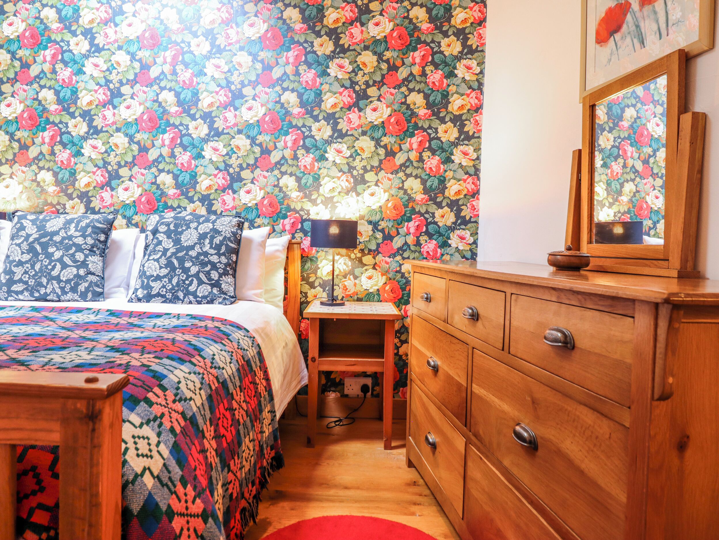 Ein Schlafzimmer mit blumigem Wandpapier, einer Holzkommode, einem Nachttisch mit Lampe und einer bettenbezogenen Tagesdecke mit Muster.