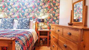 Ein Schlafzimmer mit blumigem Wandpapier, einer Holzkommode, einem Nachttisch mit Lampe und einer bettenbezogenen Tagesdecke mit Muster.