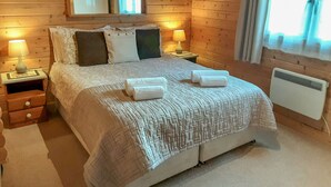 A wooden cabin bedroom with a large bed, two bedside tables with lamps, a window with curtains, and a mirror on the wall. - Pennylands Hill View Lodge (Broadway)