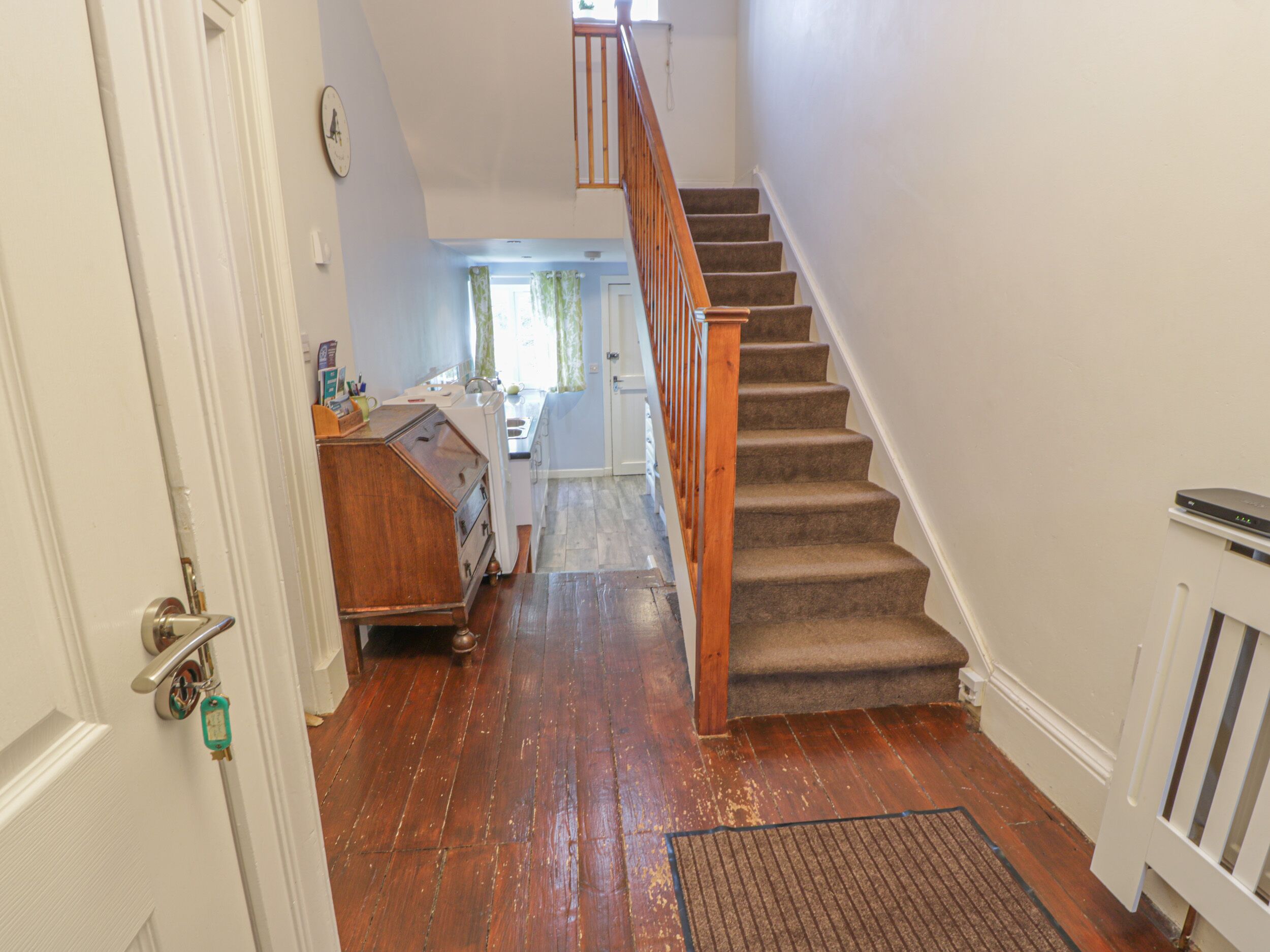 A staircase leading to a second floor with a wooden handrail and carpeted steps. A kitchen area is visible in the background with a window and curtains.