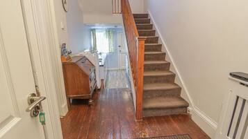 A staircase leading to a second floor with a wooden handrail and carpeted steps. A kitchen area is visible in the background with a window and curtains.