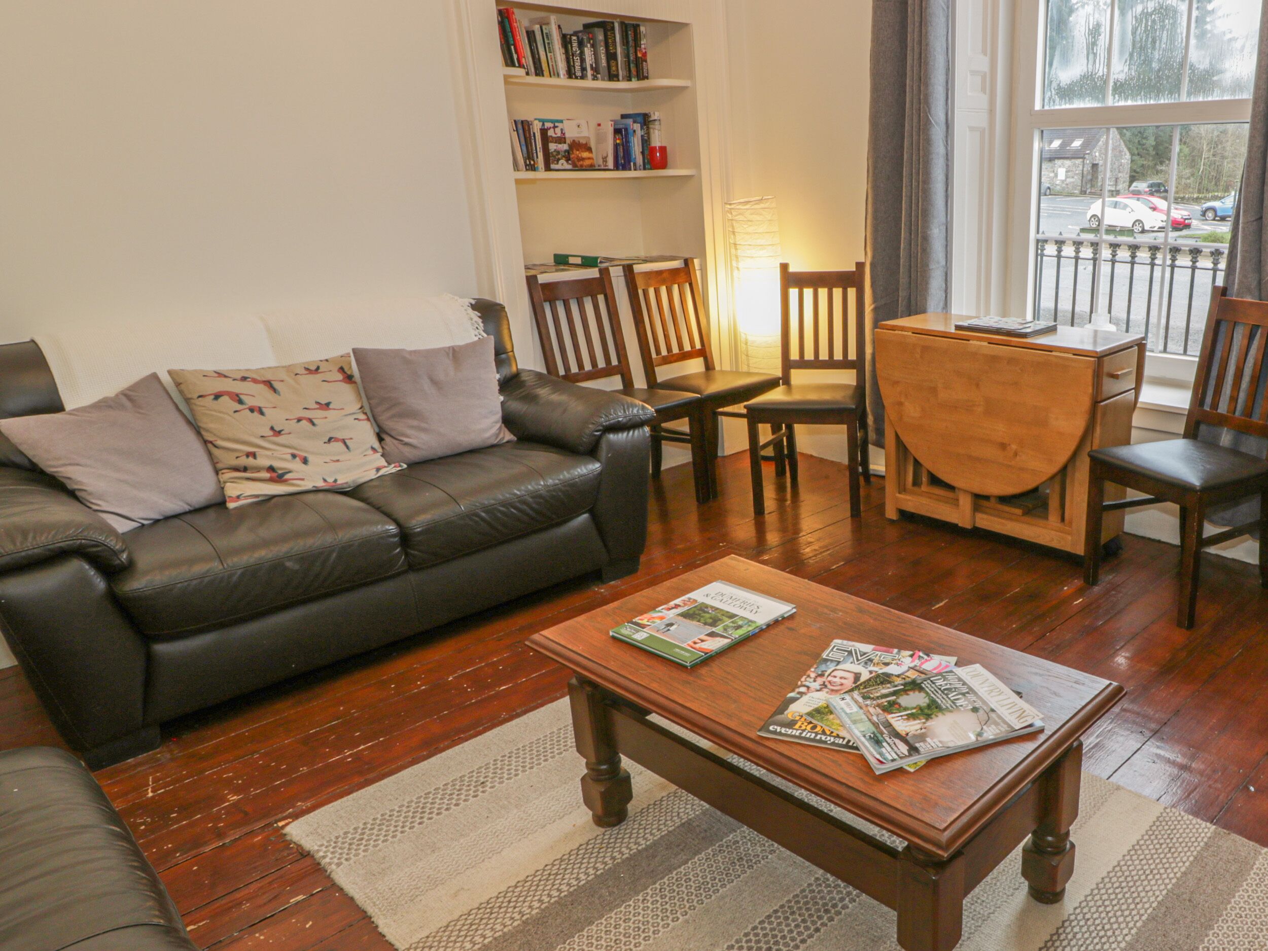 A living room with a leather sofa, wooden coffee table, and matching chairs. A bookshelf with books and a lamp is in the background.
