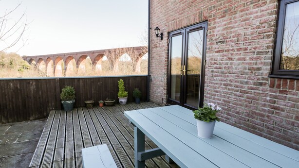 En træterrasse med et hvidt bord og bænke, krukker med planter samt en mur med et vindue og en dør.