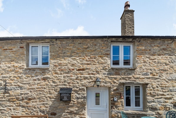 A stone building with two windows and a door, a signboard, and a chimney.