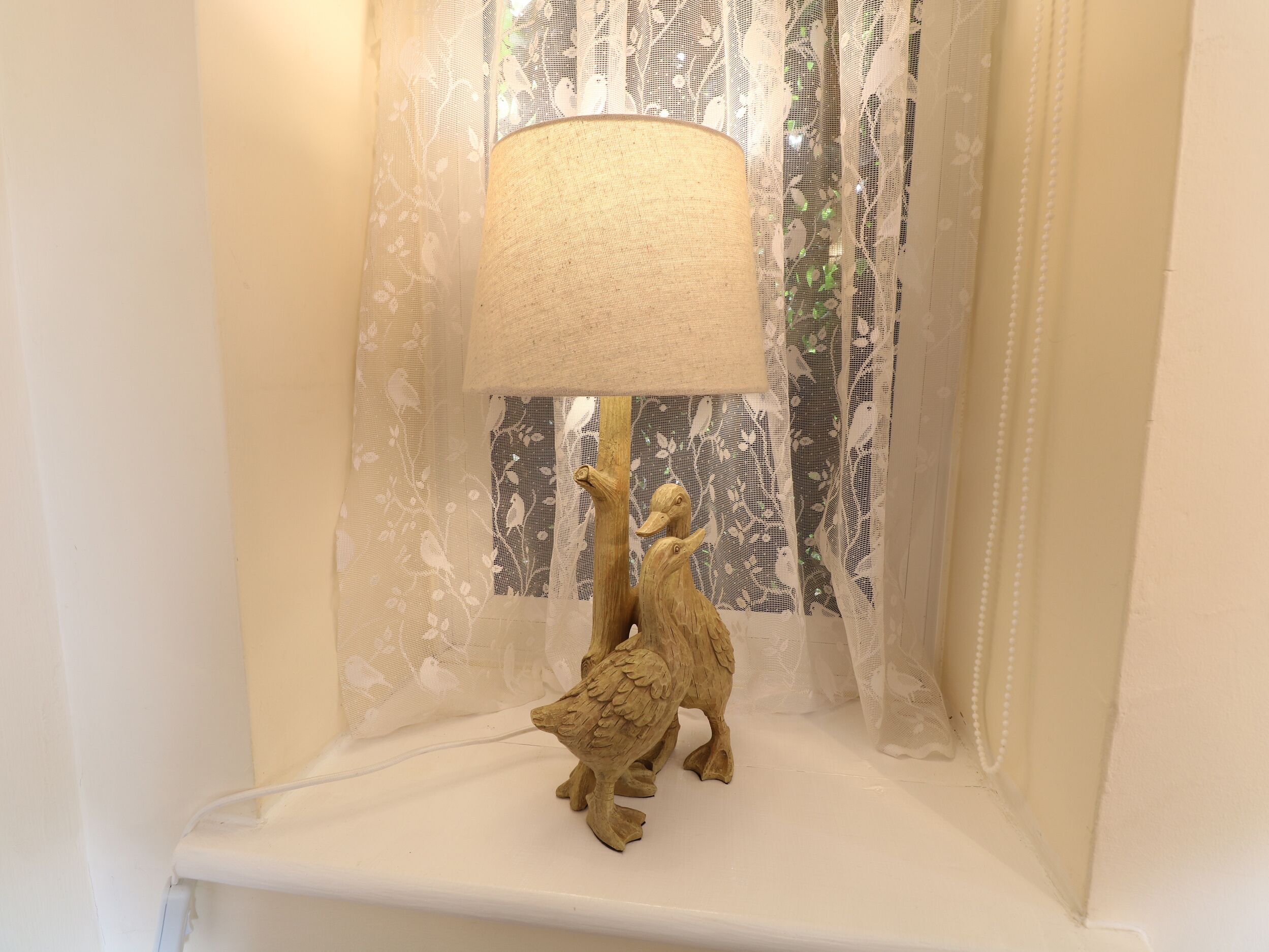 A lamp with a textured shade on a shelf, set against a window with sheer white curtains and a patterned inner curtain.
