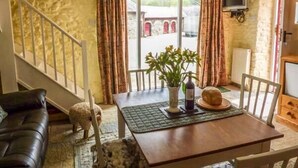 A cozy living room with a stone wall, a wooden staircase, a black leather sofa, a dining table with a vase of flowers, and a television mounted on the wall. - Crud-y-barcud (Llanwrda)
