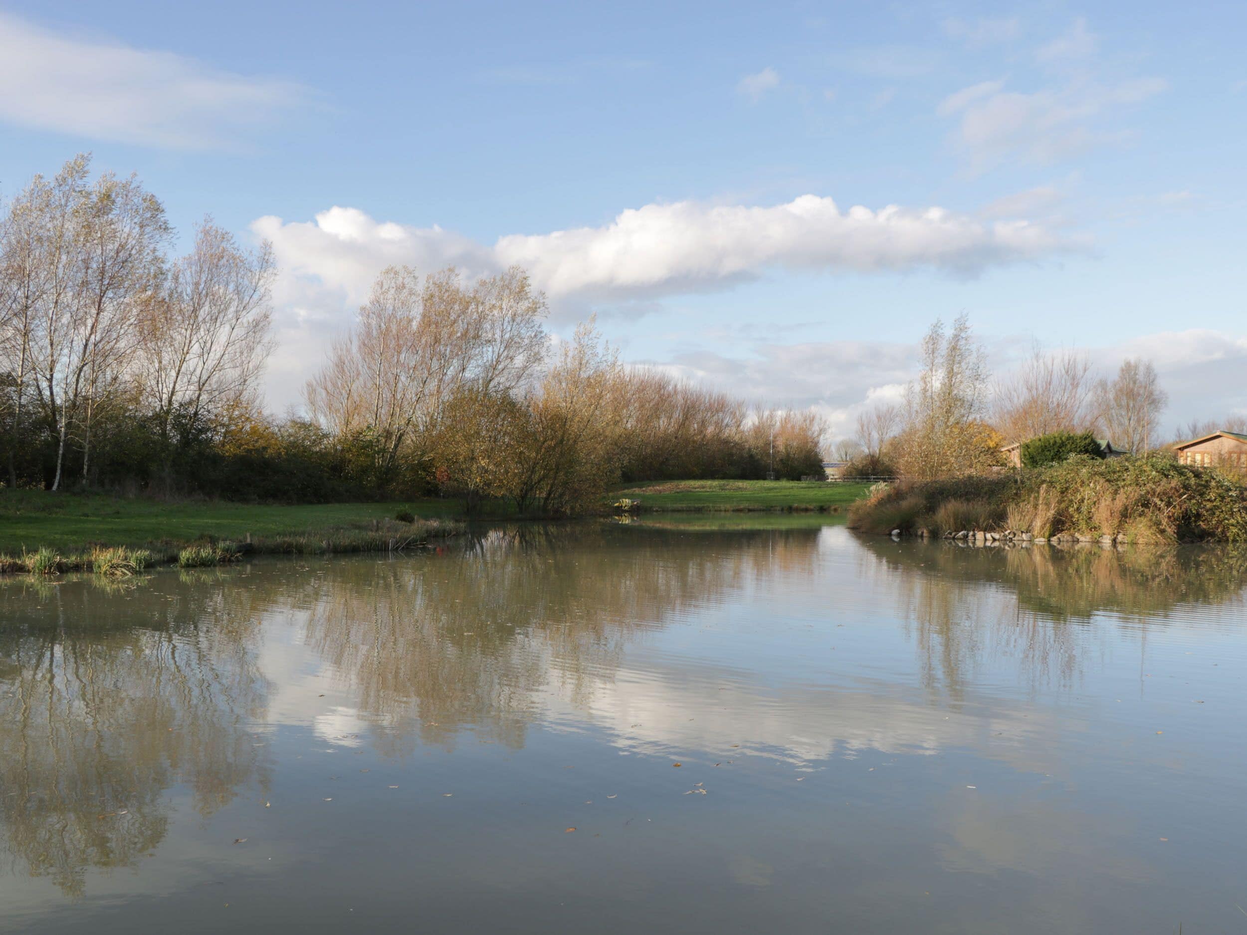 A calm river with trees reflecting on the water, surrounded by grassy areas and a building on the right.