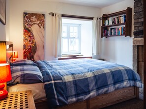 A bedroom with a bed, a nightstand, a lamp, a window with curtains, a bookshelf, and a wall painting. - Bastle House (Haltwhistle)