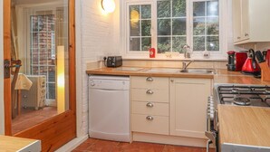 A compact kitchen with white cabinets, a built-in dishwasher, a gas stove, and a window with a view of greenery. - Beechlands Cottage (Frodsham)