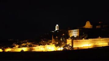 Une vue nocturne d’une ville historique, avec des fortifications illuminées et une tour imposante.