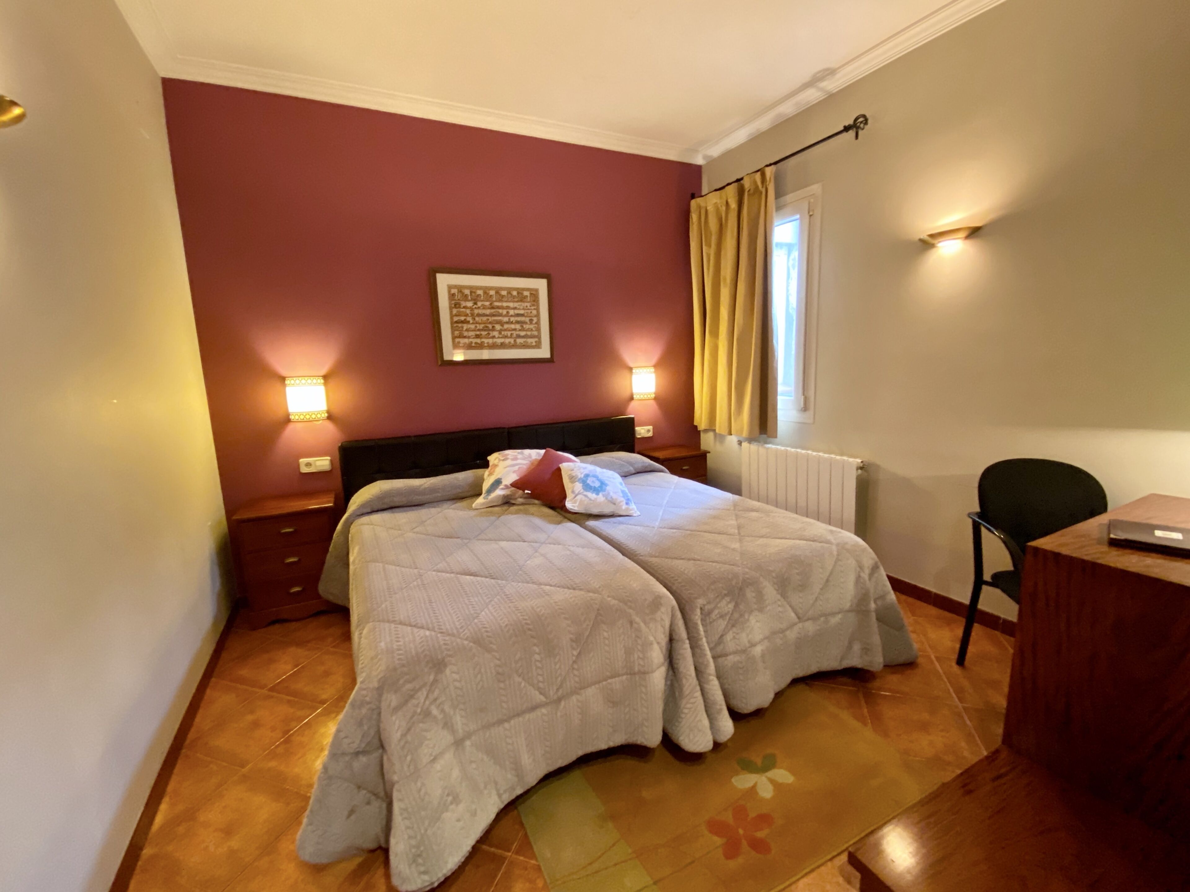 Una cama doble con colcha, dos mesitas de noche, un escritorio con silla, una ventana con cortinas e iluminación en la pared.