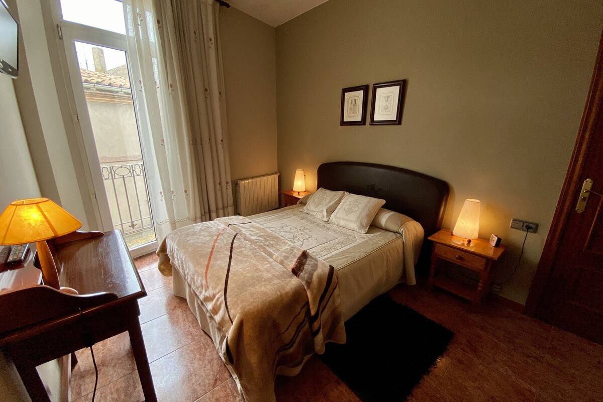 A neatly arranged bedroom with a bed, two bedside lamps, a small table, and a window with curtains.