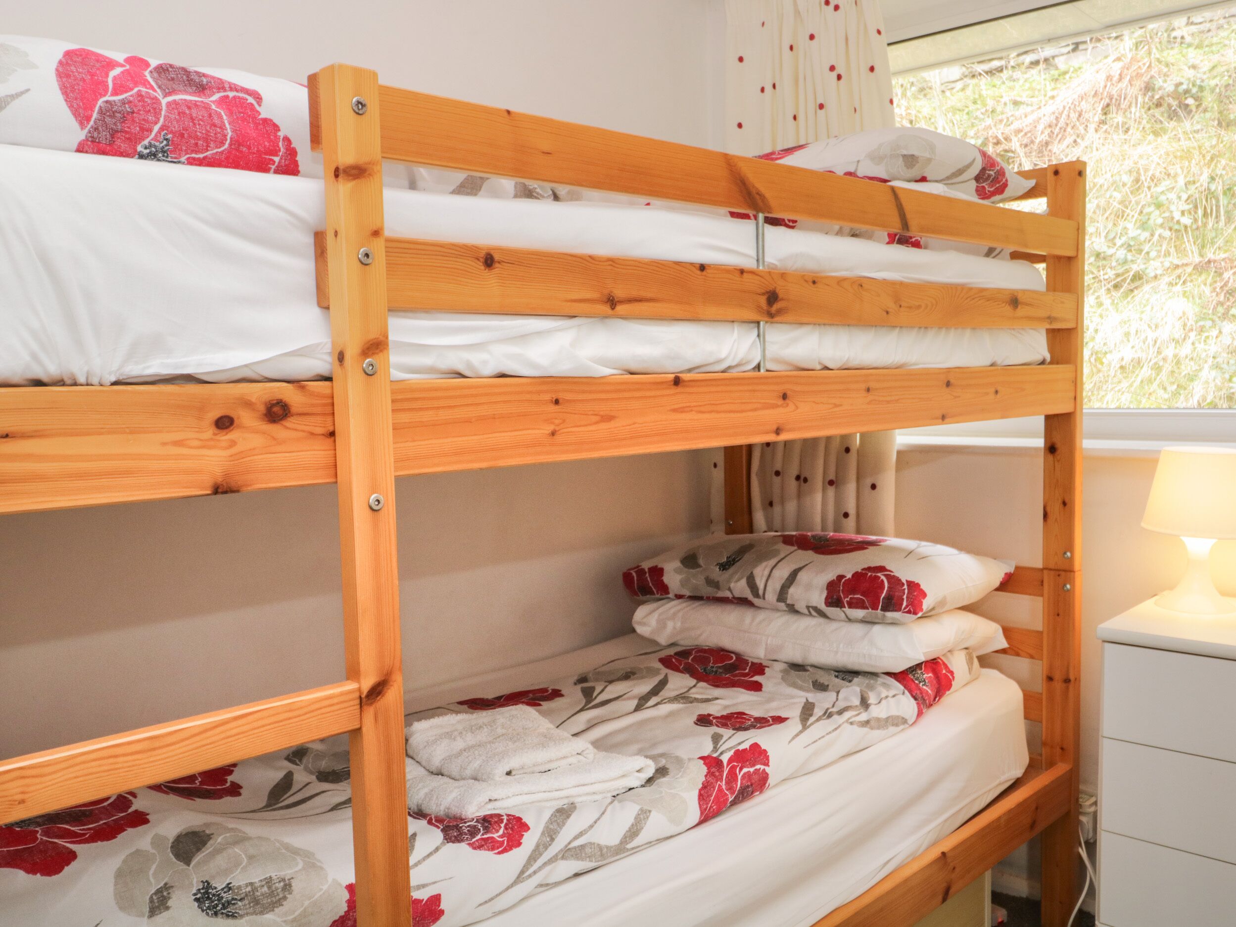 A bunk bed with white bedding and floral patterns, a wooden headboard, and a bedside table with a lamp.
