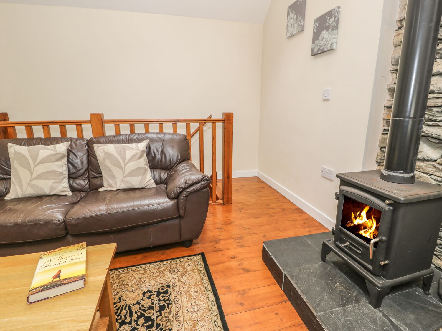 A living room with a brown leather sofa, a wooden coffee table, a book, a patterned rug, and a stone fireplace with a visible fire.
