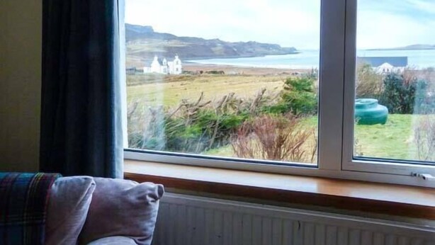 A room with a large window overlooking a coastal landscape, including a white building and a grassy field. - Amber's Cottage (Staffin)