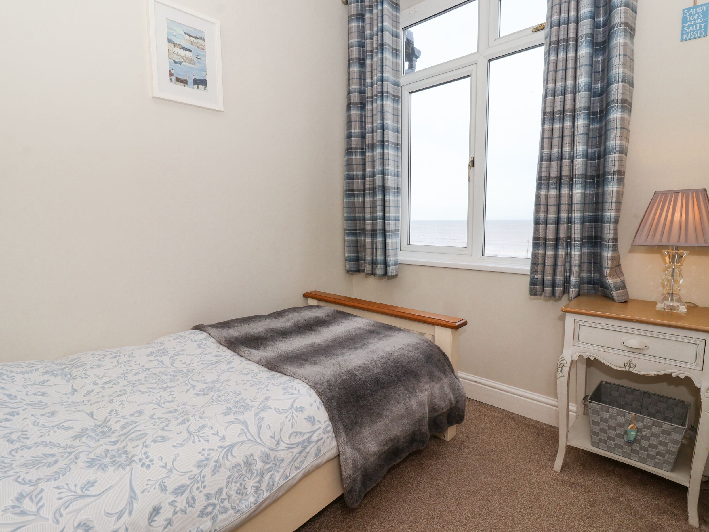 A bedroom with a bed, a window with curtains, a bedside table with a lamp, and a small wall-mounted shelf.