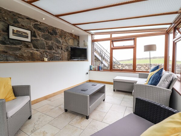 Una sala de estar acogedora con muebles de mimbre, pared de piedra, televisor y un amplio ventanal con vista a una pared de piedra y césped.
