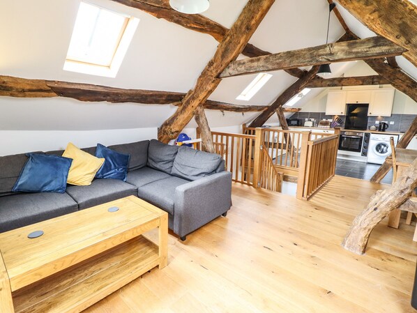 A loft-style living area with exposed wooden beams, a grey sectional sofa with blue and yellow cushions, a wooden coffee table, and a kitchen in the background. - Ysgubor Las (Dolgellau)