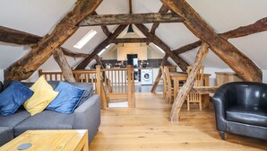 A loft-style living space with exposed wooden beams, a sofa with cushions, a coffee table, a dining area with chairs, and a kitchen in the background.