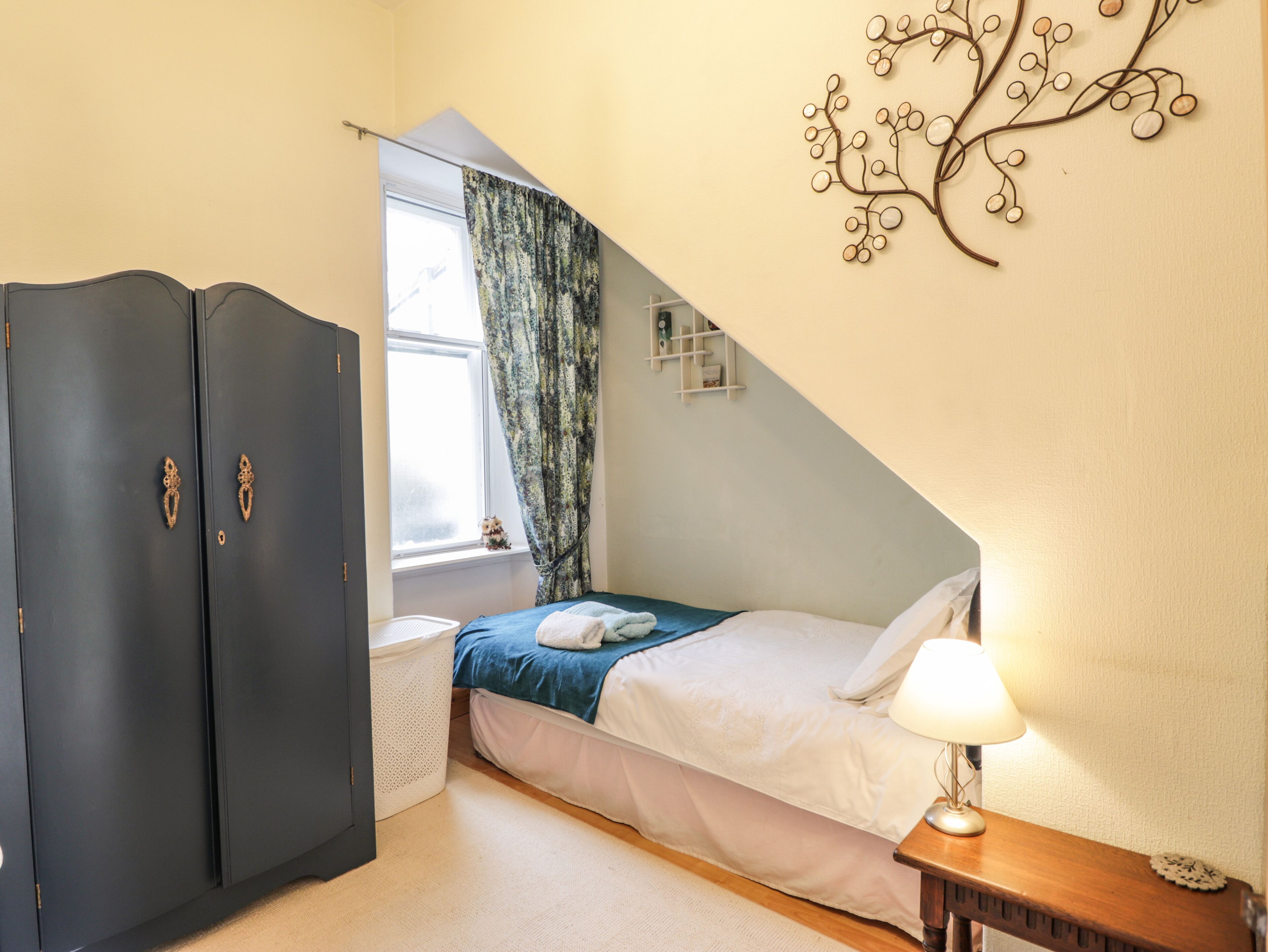 A bedroom with a bed, a nightstand, a lamp, a wardrobe, a window with curtains, and a wall decoration.