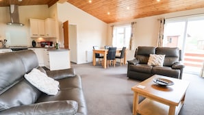 A modern living room with a leather sofa, a wooden coffee table, and a leather armchair. There is a dining area with a table and chairs, and a kitchen in the background. - Callow Lodge 5 (Shrewsbury)