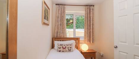 A bedroom with a single bed, a bedside table with a lamp, a window with curtains, and a door.