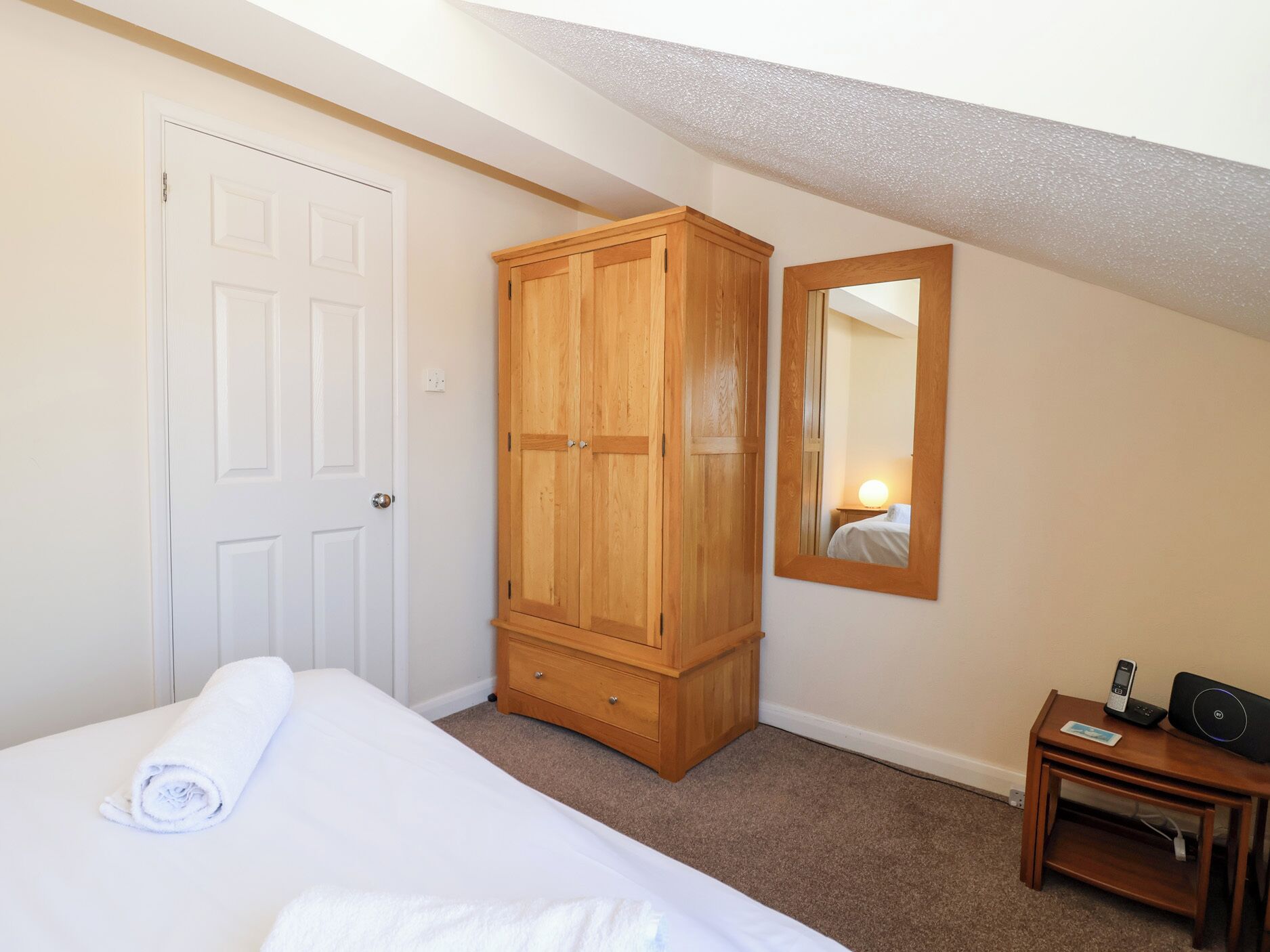 Een slaapkamer met een bed, een houten garderobe, een spiegel en een nachtkastje met een telefoon en een speaker.
