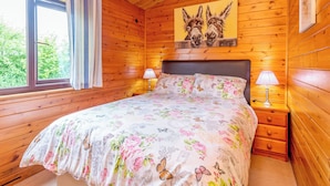 A wooden cabin bedroom with a bed, two bedside lamps, a window with curtains, and a painting of horses on the wall. - Hawthorne Lodge (Saltburn-by-the-Sea)