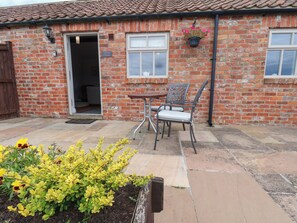 A brick building with a sign reading 'Bakehouse Cottage,' a patio with a table and chairs, and a flower bed with yellow flowers. - Blakey (Saltburn-by-the-Sea)