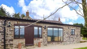 Une maison en pierre avec une grande fenêtre et une porte, une petite dépendance attenante et un arbre au premier plan.