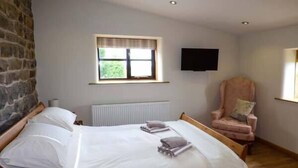 Une chambre avec deux lits, un mur en pierre, une fenêtre avec des stores, une télévision et un fauteuil rose.