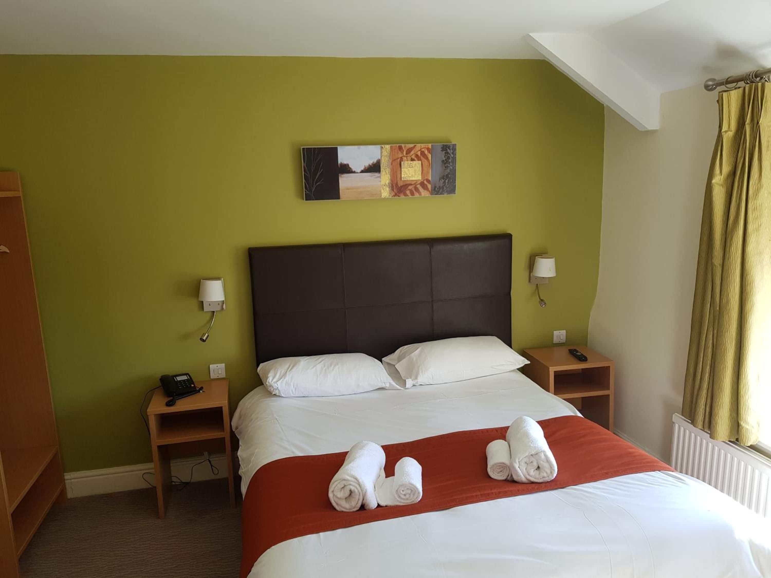 Quarto de hotel com cama, duas mesas de cabeceira, um criado-mudo, um quadro na parede e janela com cortinas.