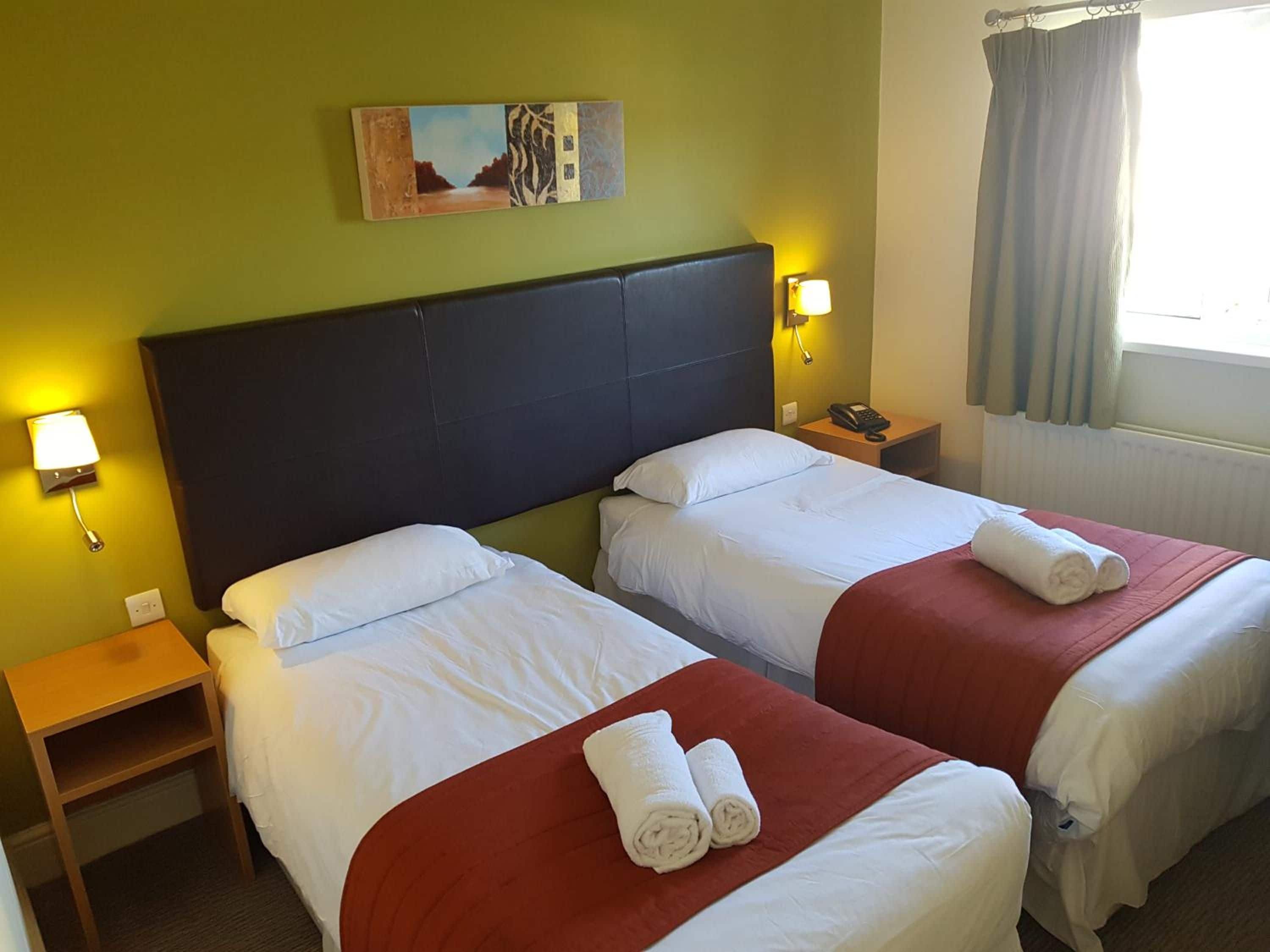 Habitación de hotel con dos camas, una mesita de noche con lámpara, un cuadro en la pared y una ventana con cortinas.