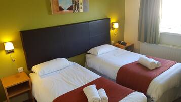 Habitación de hotel con dos camas, una mesita de noche con lámpara, un cuadro en la pared y una ventana con cortinas.