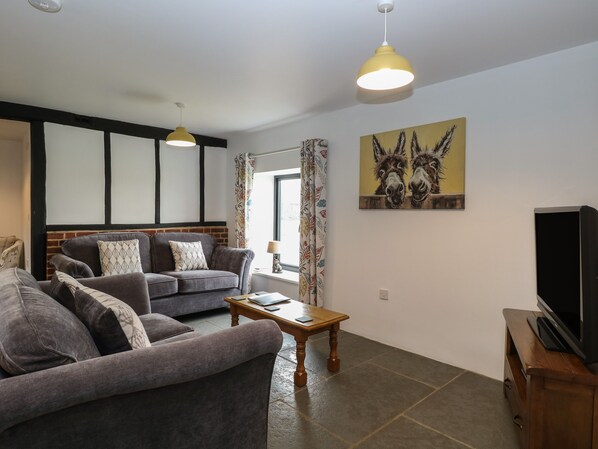 A living room with a grey sofa set, a wooden coffee table, a TV on a wooden stand, and a painting of donkeys on the wall. - Tricker's Cottage (Norwich)