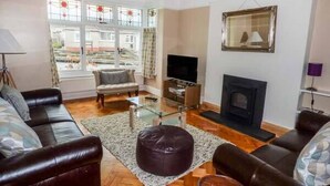 A living room with a sofa, ottoman, TV, fireplace, and large windows with curtains. - Pen Hill (Llantwit Major)