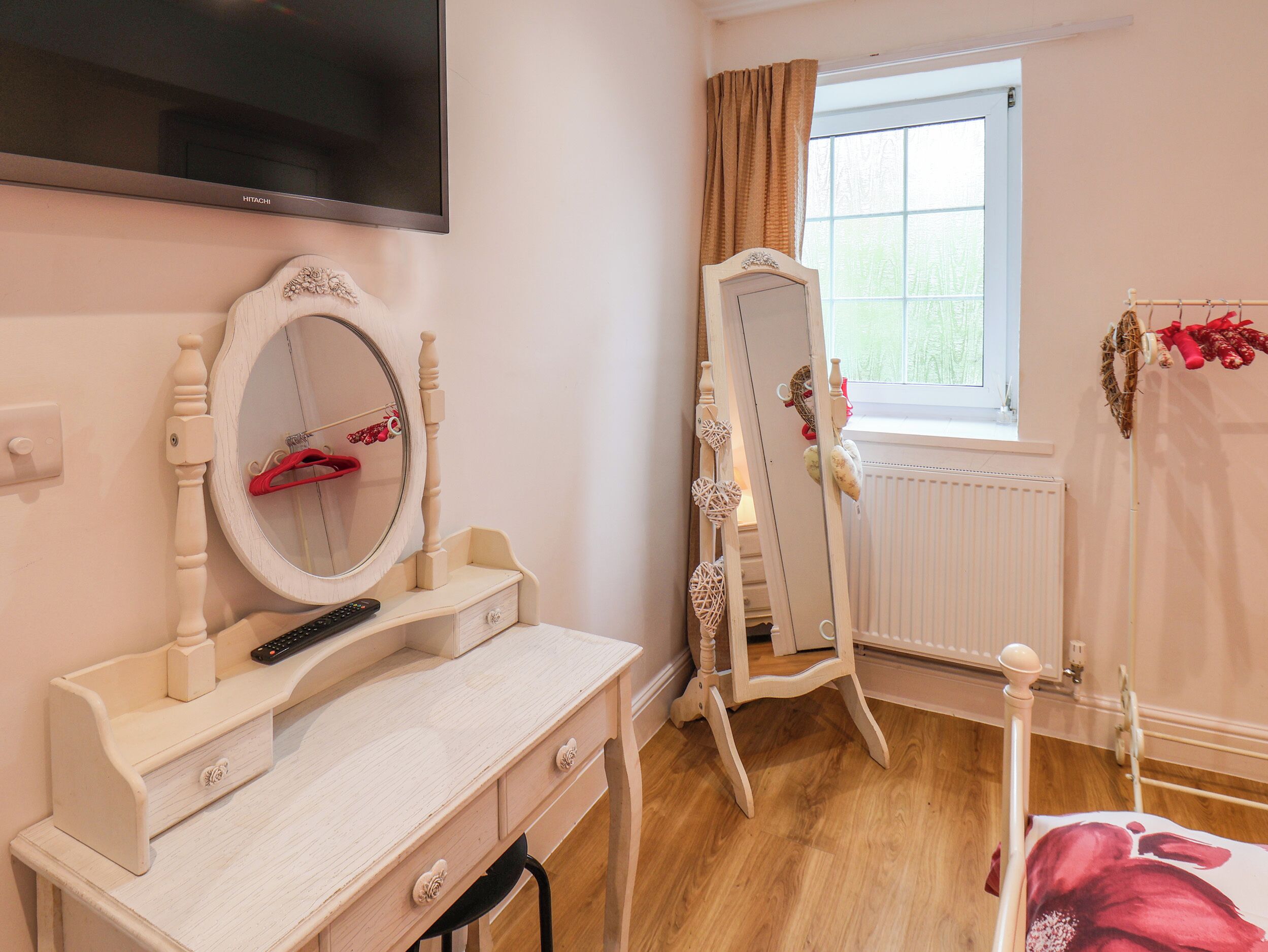 Ein Zimmer mit an der Wand montiertem Fernseher, einem Schminktisch mit Spiegel, einem Schmuckkästchen, einem Frisiertisch mit Spiegel, einem Heizkörper und einem Fenster mit Vorhängen.