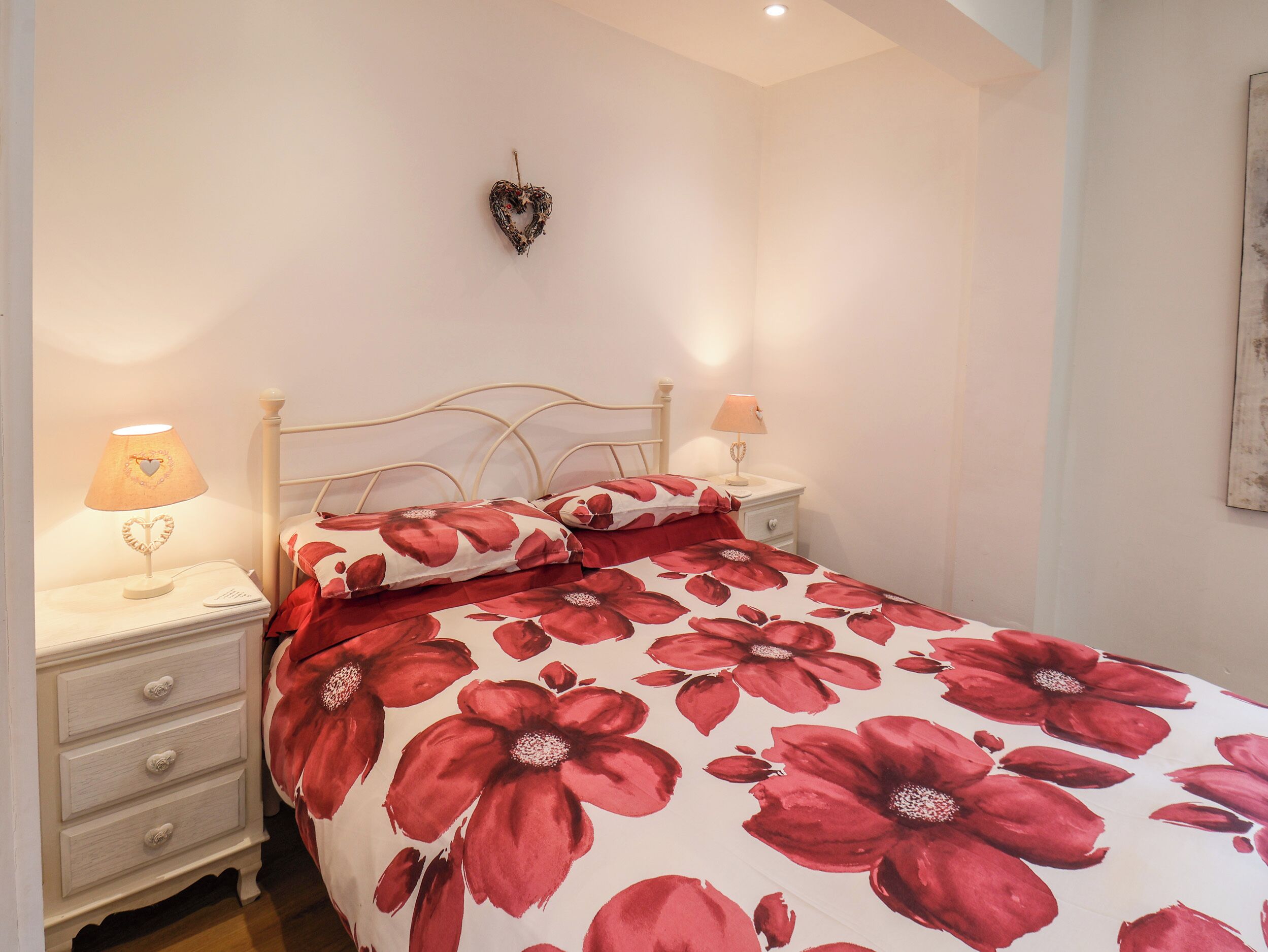 A bedroom with a bed, two bedside tables, a wall-mounted heart decoration, and a nightstand with a lamp.
