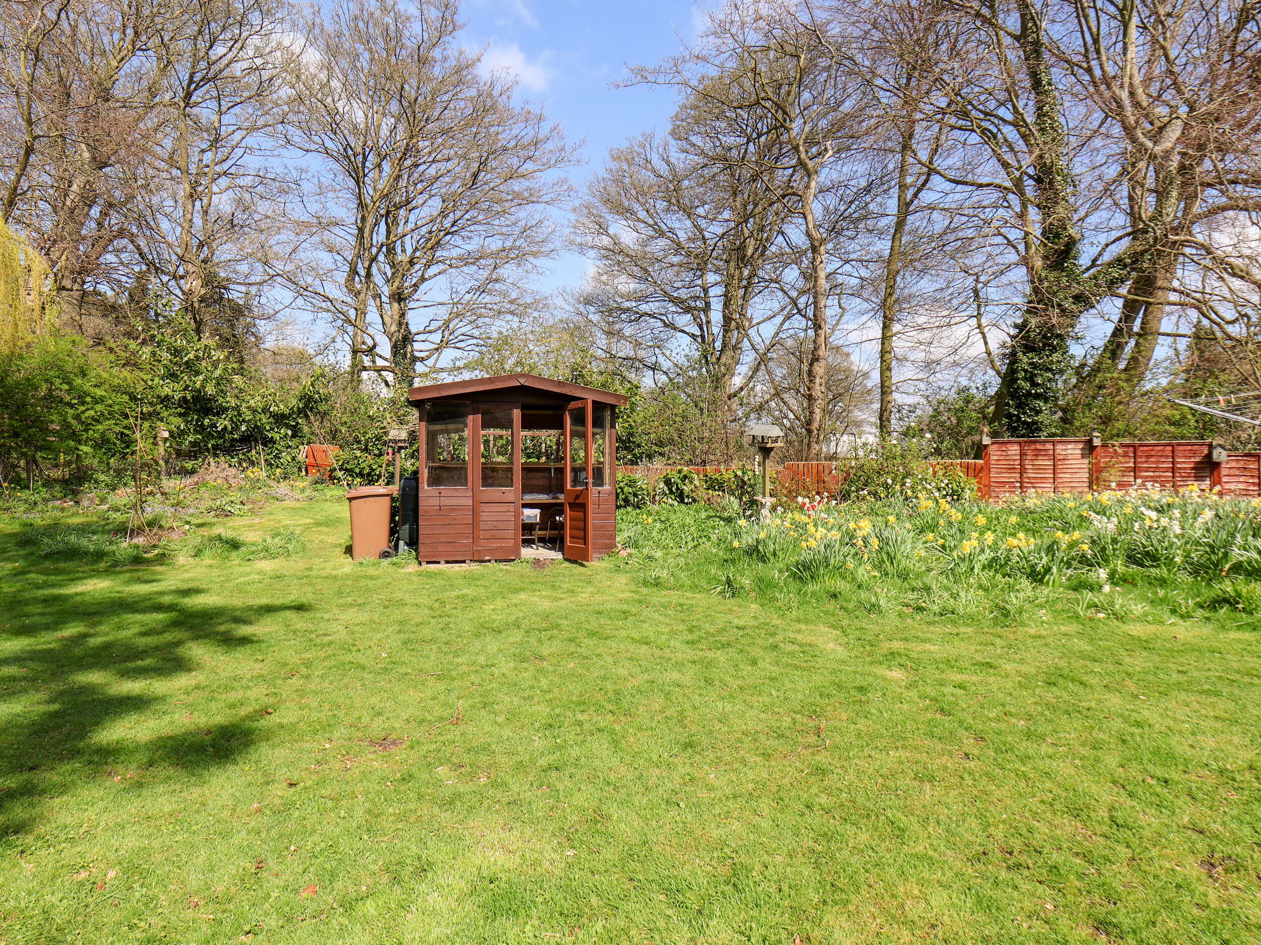 A wooden gazebo with a bench, surrounded by a well-maintained garden with various plants and flowers.