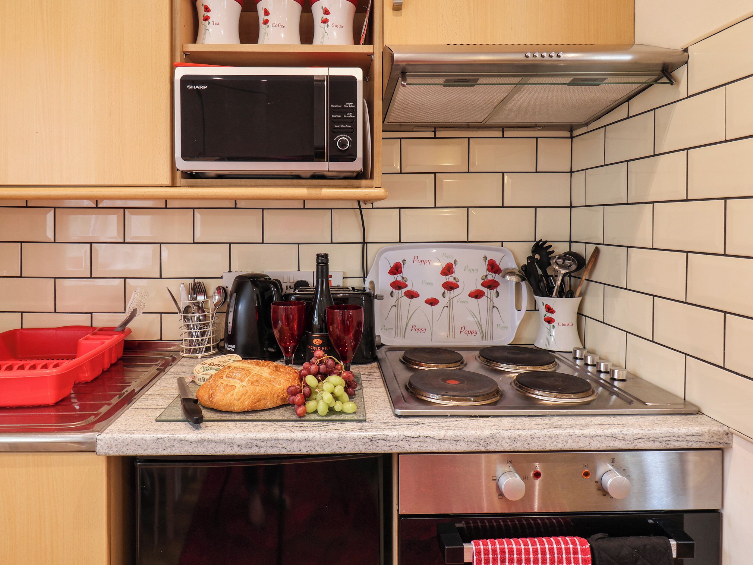A kitchen with a microwave, toaster, and stove. There are red and white mugs on a shelf, a red dish rack, and a basket of bread and grapes on the counter.