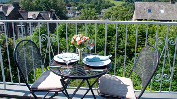 En balkon med et bord dækket til to, en blomstervase og en smuk udsigt over en bakke og bygninger i baggrunden.