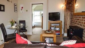 A living room with a leather sofa, a coffee table, a TV on a wooden stand, and a brick wall. - Bluebird Loft (Coniston)
