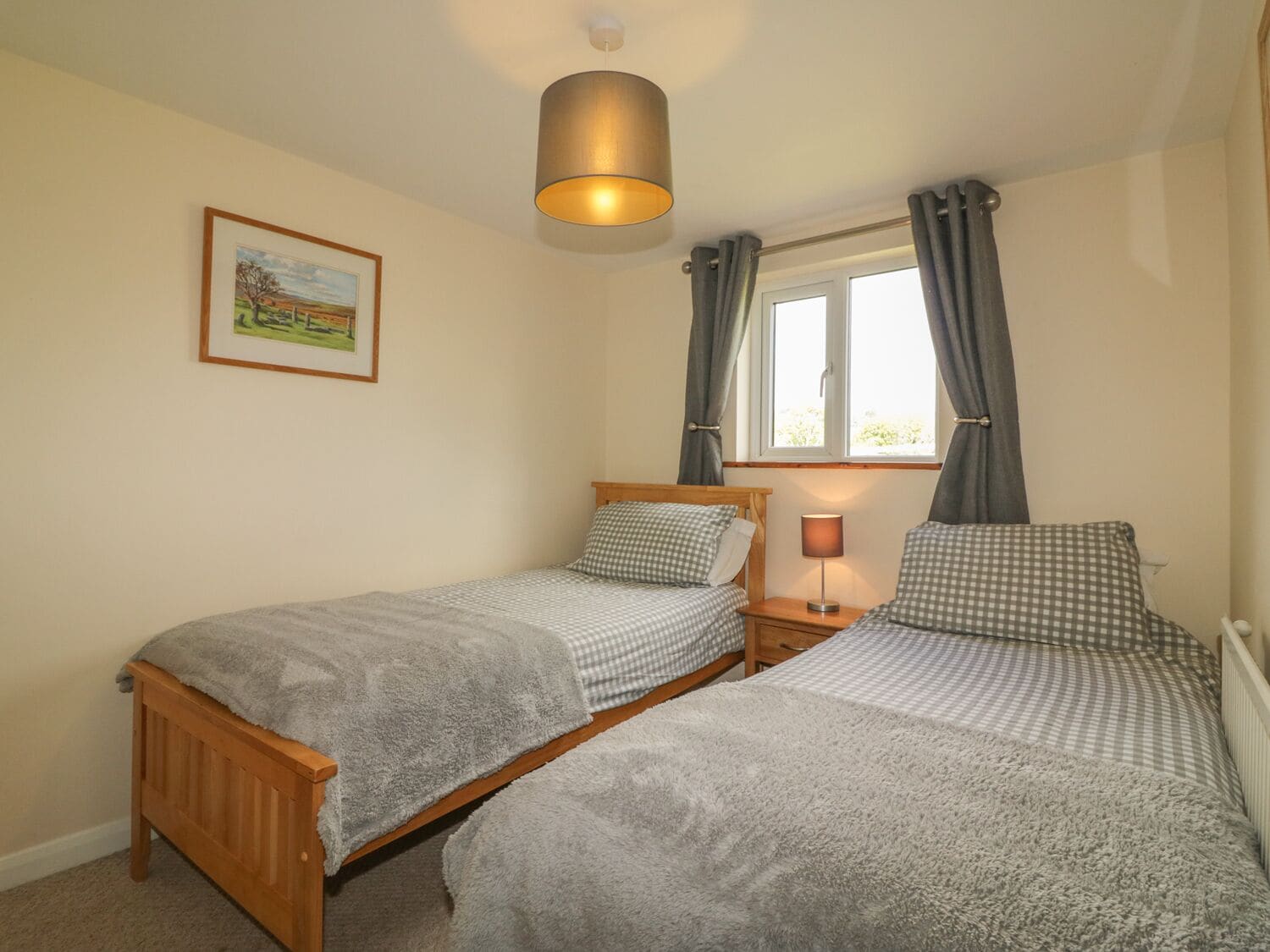 A bedroom with two single beds, a bedside table with a lamp, a window with curtains, and a framed picture on the wall.